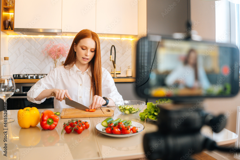 Medium shot portrait of attractive redhead woman vlogger shooting video food blog about cooking on camera of mobile phone, sitting at kitchen with modern light interior, selective focus.
