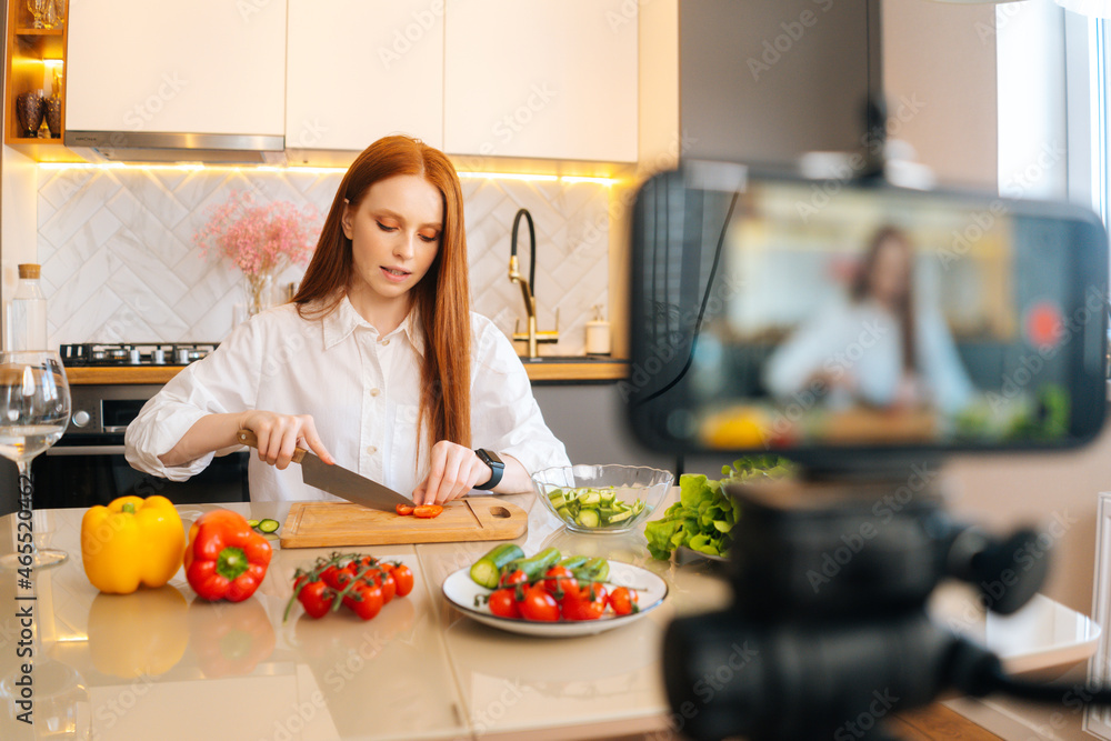 Medium shot portrait of attractive redhead woman vlogger shooting video food blog about cooking on camera of mobile phone, sitting at kitchen with modern light interior, selective focus. Medium shot portrait of attractive redhead woman vlogger shooting video food blog about cooking on camera of mobile phone, sitting at kitchen with modern light interior, selective focus.