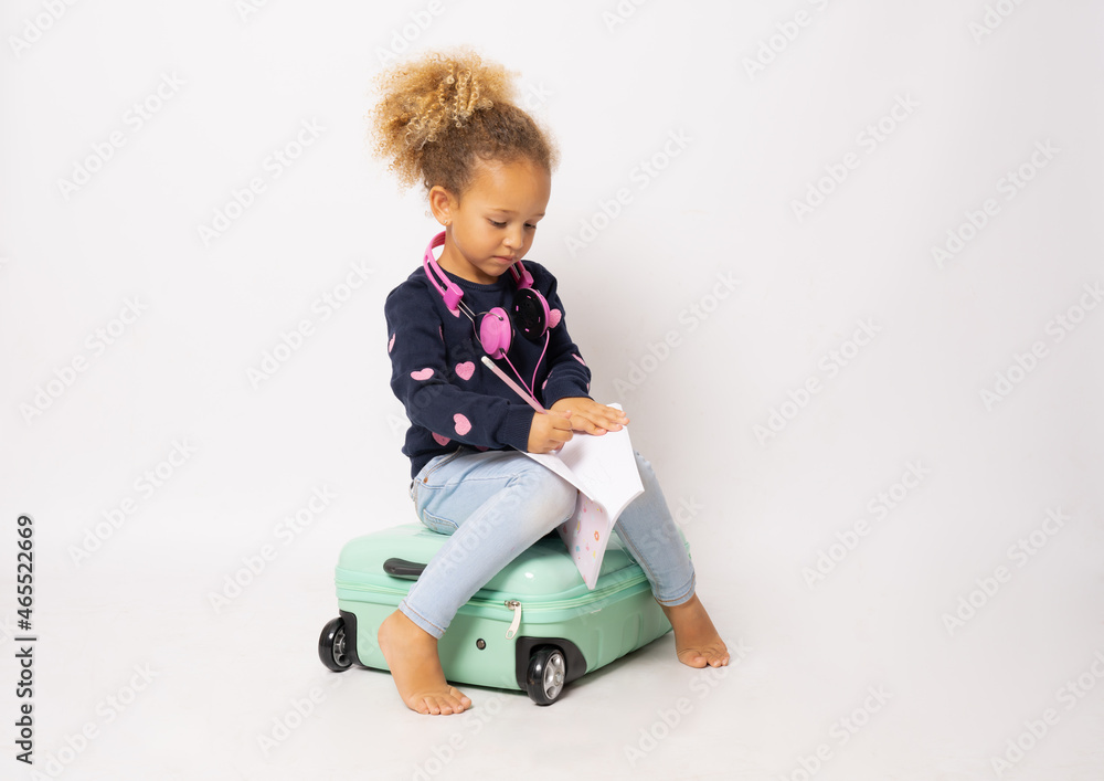 Beautiful little girl sitting on a suitcase and holding a notebook isolated over white background. Beautiful little girl sitting on a suitcase and holding a notebook isolated over white background.