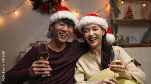 Happy young sweet couple in santa hat making video call and talking to camera greeting family on Christmas and New Year holidays. two female and male lovers sitting on couch with glasses of wine