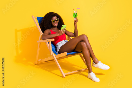 Portrait of attractive cheerful girl drinkinhg mojito sitting in chair using device isolated over bright yellow color background