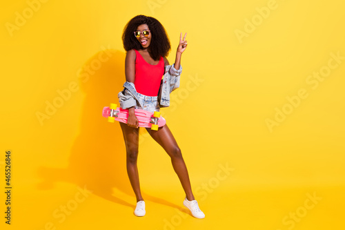 Full length body size photo of girl keeping skate board showing v-sign gesture isolated on vibrant yellow color background