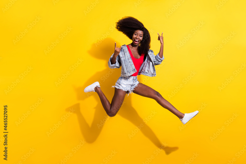 Full length body size photo of girl jumping showing heavy metal sign laughing isolated bright yellow color background Full length body size photo of girl jumping showing heavy metal sign laughing isolated bright yellow color background
