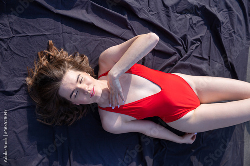 Young caucasian woman in red swimsuit lying on black cloth closed her eyes from the sun