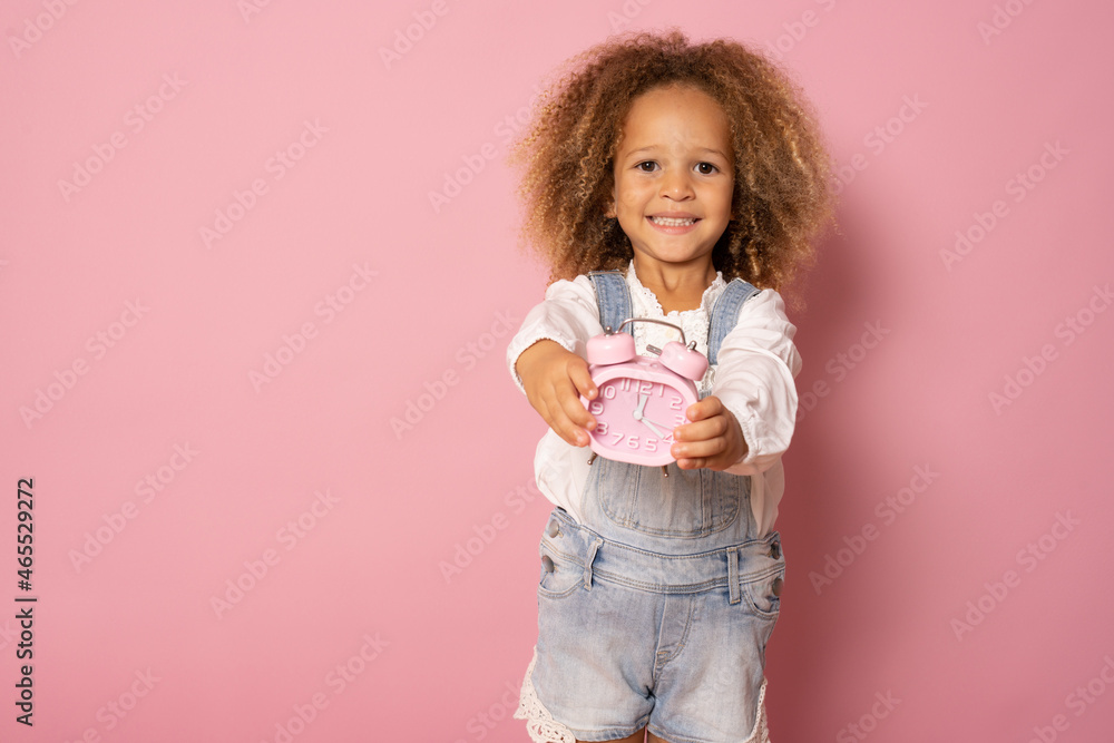 Adorable little kid girl in casual clothing smiling and holding alarm clock isolated over pink background Adorable little kid girl in casual clothing smiling and holding alarm clock isolated over pink background