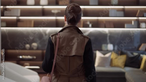 Back view concentrated wealthy woman entering furniture store showroom looking around and leaving. Live camera follows elegant slim beautiful rich Caucasian lady walking in shop choosing furnishings
