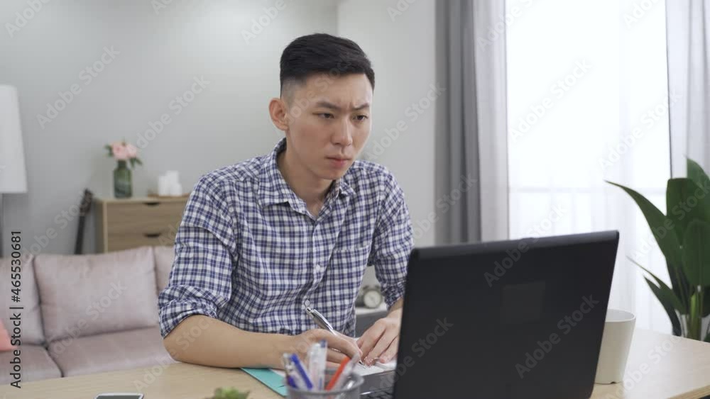 asian manager is speaking with hand gestures and taking down notes during an online briefing with laptop in the living room at home. asian manager is speaking with hand gestures and taking down notes during an online briefing with laptop in the living room at home.