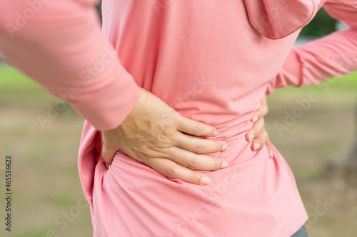 Young woman in sports outfits pink injured her backache during exercise in the park. Upper section of sports girl suffering from backache while sitting at workout. Accident from exercise concept.