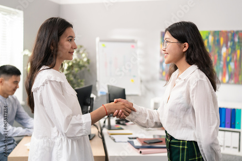 Two women elegantly dressed are standing in middle of corporate office, they are smiling at each other shaking hands in friendly manner, greeting, saying goodbye, getting acquainted, congratulating © ABCreative