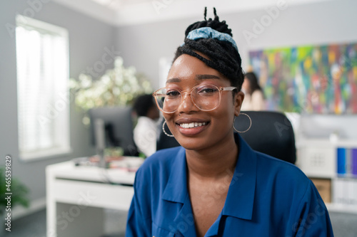 Attractive lady with dark complexion, dreadlocks tied in a bun wearing a blue shirt and glasses smiles looking into the camera, secretary, office job, corporation, corporate room, customer service
