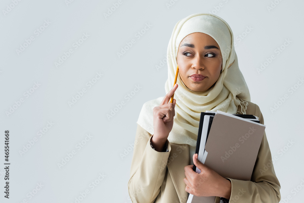 pensive muslim woman in hijab holding pencil and folders isolated on ...