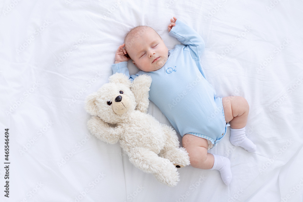 baby boy sleeps on the bed lying on his back with a soft toy bear in blue pajamas holding his hands up, healthy newborn sleep baby boy sleeps on the bed lying on his back with a soft toy bear in blue pajamas holding his hands up, healthy newborn sleep