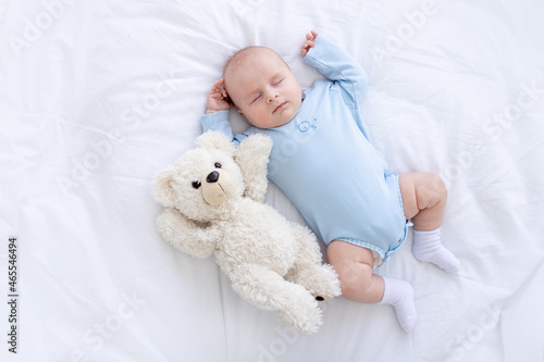 baby boy sleeps on the bed lying on his back with a soft toy bear in blue pajamas holding his hands up, healthy newborn sleep