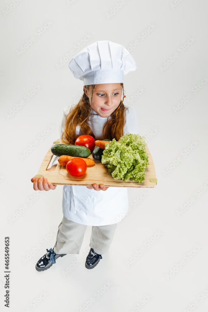 High angle view of little cute girl in white cook uniform and huge chef's hat holding tray with healthy vegetables isolated on white studio background. High angle view of little cute girl in white cook uniform and huge chef's hat holding tray with healthy vegetables isolated on white studio background.