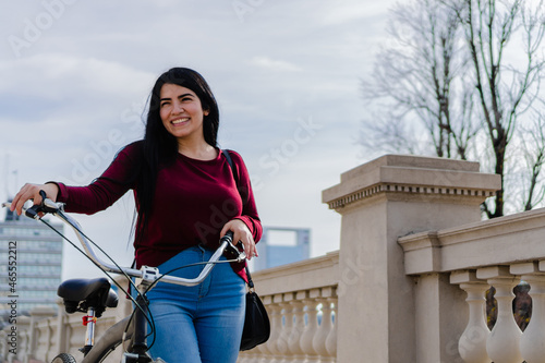 young latin tourist woman looking away, with her bicycle in her hand, smiling, walking in the city, on a stroll.