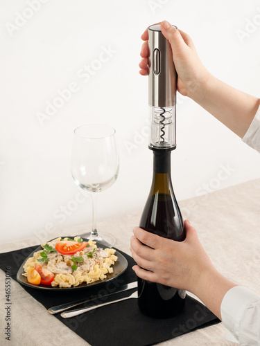 A woman's hand opens a bottle of wine with an electric corkscrew made of gray metal. Next to it is a plate of pasta, a glass and cutlery.