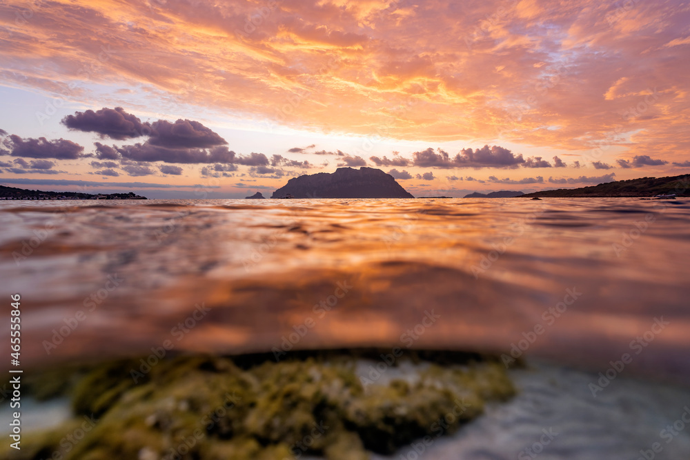 (Selective focus) Split shot, over under water surface. Defocused waves in the foreground with Tavolara Island on the surface during a dramatic sunrise. Porto Istana, Sardinia, Italy.