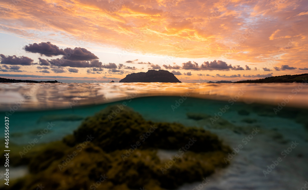 Fototapeta premium (Selective focus) Split shot, over under water surface. Defocused waves in the foreground with Tavolara Island on the surface during a dramatic sunrise. Porto Istana, Sardinia, Italy.