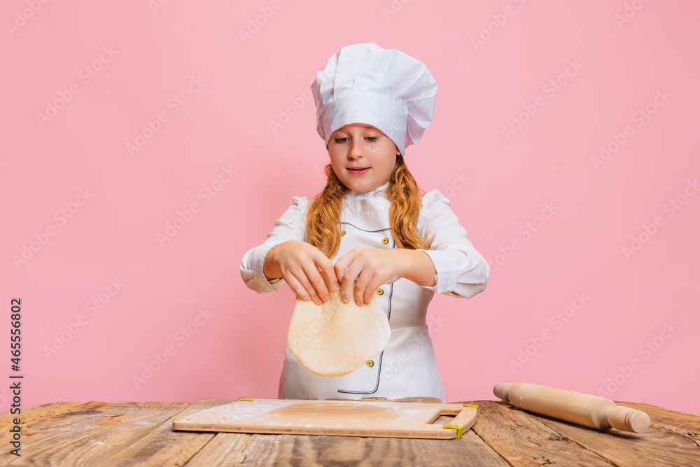 Portrait of little beautiful girl in white cook uniform and huge chef's hat at kids kitchen isolated on pink studio background. Portrait of little beautiful girl in white cook uniform and huge chef's hat at kids kitchen isolated on pink studio background.