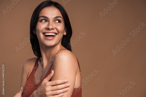 Laughing vivacious young woman with a beaming smile embracing herself with her hands while posing over brown background. Happy woman with naked shoulders laughing and looking away
