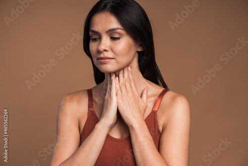 Portrait of ill woman with brown hair standing and touching her neck while frowning from pain, suffering sore throat, flu symptom. Indoor studio shot isolated on brown background