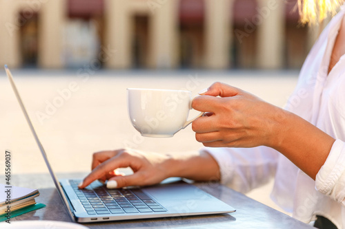 Close up of woman hands holding a cup of coffee and typing on laptop outdoors at cafe table. Businesswoman or student browsing internet and drinking morning coffee.