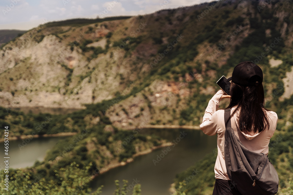 woman in a cap with a backpack takes pictures of the mountains and the river on the phone woman in a cap with a backpack takes pictures of the mountains and the river on the phone