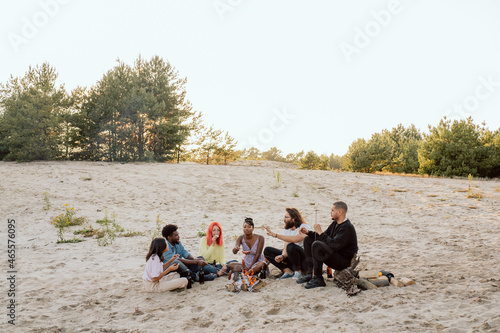 Socializing students get to know each other on campfire, different skin colors, nationalities, they talk, joke, laugh, sit on the sand near fire, fry sausages on sticks, drink beer, view from afar