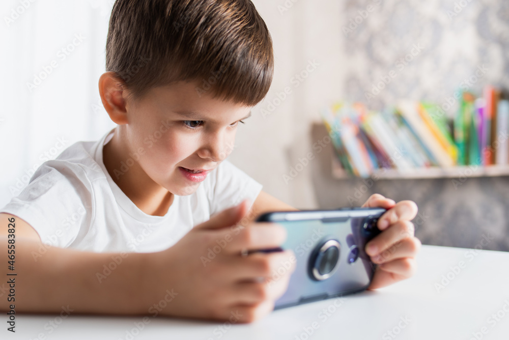 A cheerful child in a white T-shirt plays games on the phone at home. Happy boy looks at his smartphone. A cheerful child in a white T-shirt plays games on the phone at home. Happy boy looks at his smartphone.