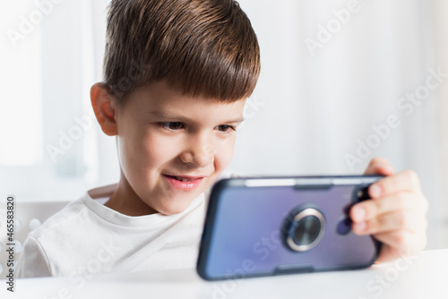 A little boy in a white T-shirt plays games on the phone at home. A happy child looks at his smartphone.