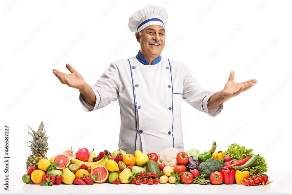 Cheerful mature chef smiling and gesturing with hands behind a pile of fruits and vegetables Cheerful mature chef smiling and gesturing with hands behind a pile of fruits and vegetables