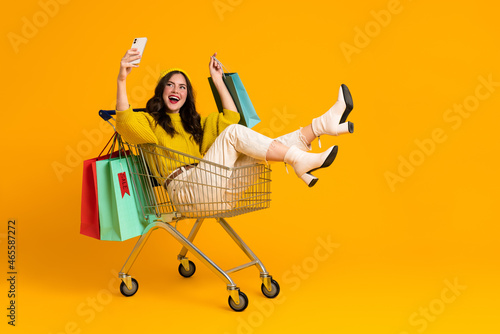 Fototapeta White excited woman taking selfie on cellphone in shopping cart