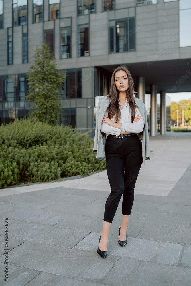 Portrait of tired corporate executive in daily makeup woman dressed in white shirt with jacket thrown over back arms crossed over chest stand outside in front of a modern corporate office building Portrait of tired corporate executive in daily makeup woman dressed in white shirt with jacket thrown over back arms crossed over chest stand outside in front of a modern corporate office building