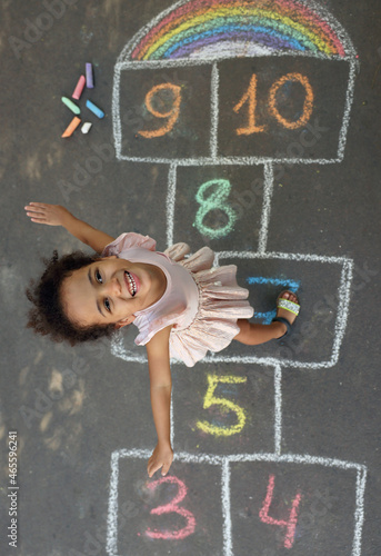 Little African American girl and colorful hopscotch drawn with chalk on asphalt outdoors, top view. Happy childhood photo