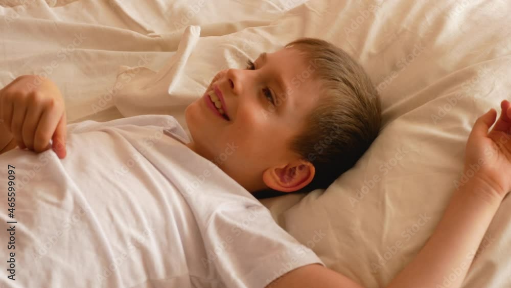 Close-up of a Caucasian boy 7-9 years old wakes up in the daylight with a good mood. The child wakes up and stretches out his arms while lying on the bed. Good morning I woke up. Selective focus Close-up of a Caucasian boy 7-9 years old wakes up in the daylight with a good mood. The child wakes up and stretches out his arms while lying on the bed. Good morning I woke up. Selective focus