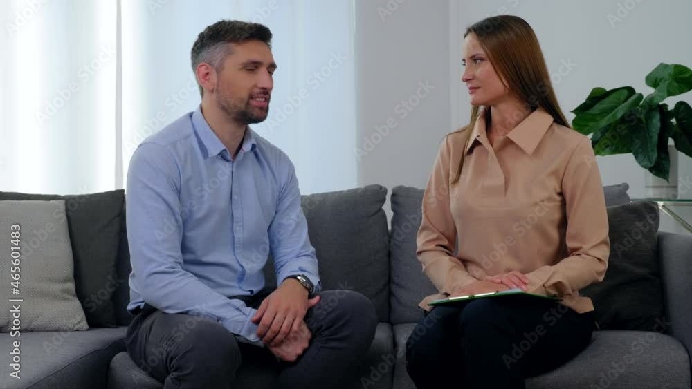 Woman psychologist and man patient sitting on couch talking and turn to camera with smile after having passed positive psychotherapy session in clinic office. Mental health problems after covid-19 Woman psychologist and man patient sitting on couch talking and turn to camera with smile after having passed positive psychotherapy session in clinic office. Mental health problems after covid-19