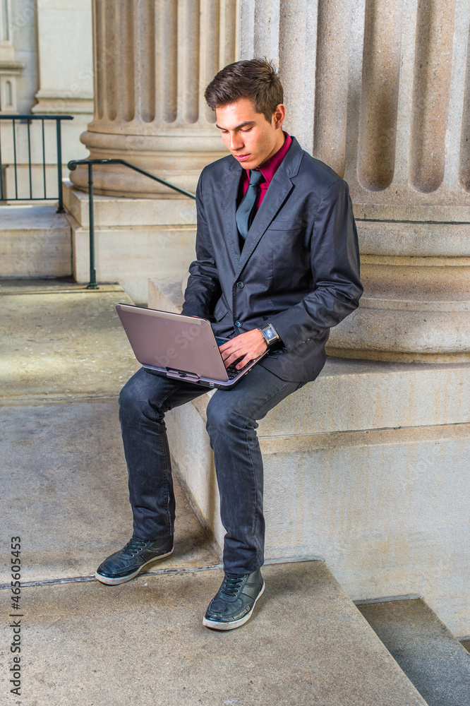 Dressing in a red undershirt, a black blazer, jeans, a black tie, a young college student is working on a laptop computer outside an office on campus.. Dressing in a red undershirt, a black blazer, jeans, a black tie, a young college student is working on a laptop computer outside an office on campus..