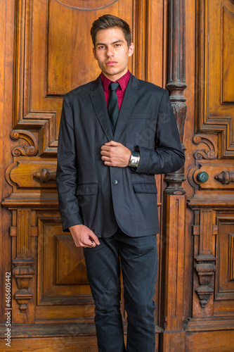 Dressing in a red undershirt, a black blazer, black jeans and a black tie, a young handsome college student is standing in the front of old fashion style door, looking forward.