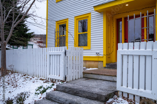 The corner of a white clapboard wood house with multiple double hung windows with vibrant yellow trim, and a red door. There's a white wooden picket fence in the front of the building with snow.