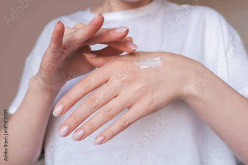 Female hands with natural pink manicure applying moisturizing cream or lotion on beige background. Bodycare  skin protection concept. Woman in white uses beauty product. Spa procedure closeup. 