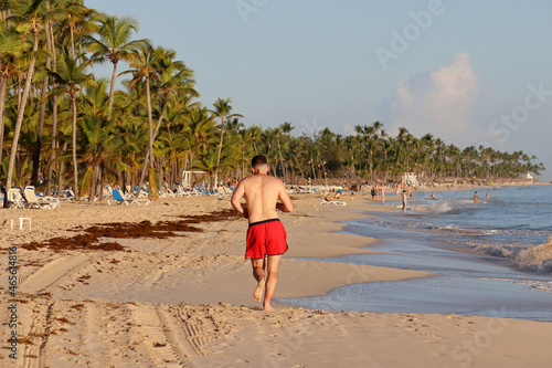 Barefoot man in red shorts running by the sand on a tropical beach on coconut palm trees background. Workout on a beach at morning, healthy lifestyle