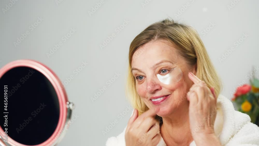 Old woman using eyes patches close-up, looking in mirror. Face skin care, beauty procedure, morning routine in bathroom. Cosmetology concept, cosmetic products. Old woman using eyes patches close-up, looking in mirror. Face skin care, beauty procedure, morning routine in bathroom. Cosmetology concept, cosmetic products.