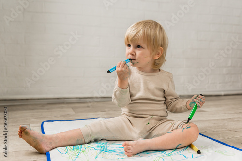 A little cheerful girl in pajamas is sitting on the floor and drawing with water markers on special oilcloth. The face and clothes are stained with paint. Tastes the paint