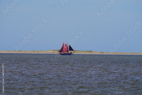 an old wooden sailing boat with two masts and red sails sails close to the coast with sand dunes and a sandy beach on which people go for a walk