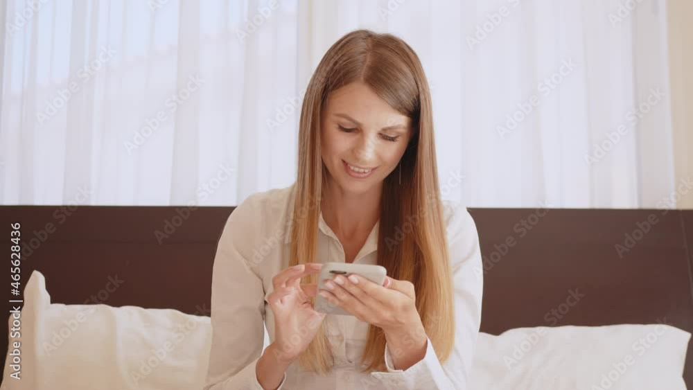 Charming young woman with long hair surfing internet on modern smartphone while resting at hotel. Happy caucasian lady sitting on bed and using cell phone. Charming young woman with long hair surfing internet on modern smartphone while resting at hotel. Happy caucasian lady sitting on bed and using cell phone.