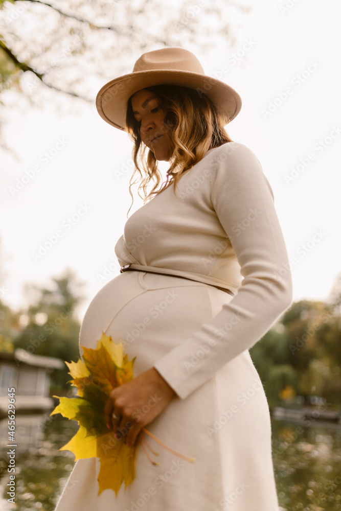 Pregnant girl in a hat and a white dress in autumn at sunset in the park