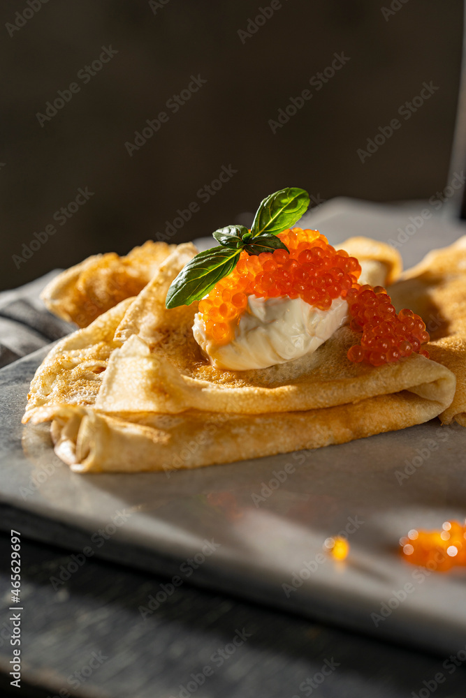 Crepes (big thin pancakes) with red caviar, and cream cheese or sour cream with basil leaf on dark background. Beautiful light, selective focus, copy space. Tasty breakfast, dark mood photo concept.