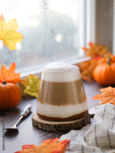 A cup of hot coffee layer caramel latte and mouse foam milk on a table near the window with autumn atmosphere with yellow maple leaves and Jack-Be-Little pumpkin in background