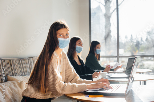 A group of masked girls keep a social distance in a cafe when working on laptops.