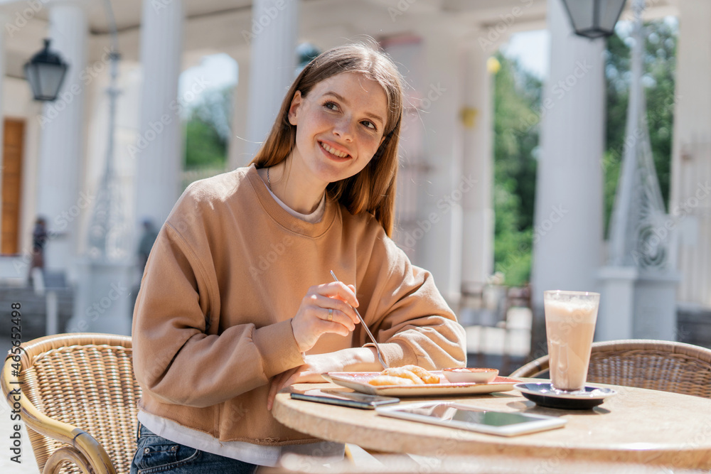 A female student is sitting in a summer cafe. Freelancer surf the internet information. Smiles and prints a message on a social network. Millennial communicates with friends online. A female student is sitting in a summer cafe. Freelancer surf the internet information. Smiles and prints a message on a social network. Millennial communicates with friends online.